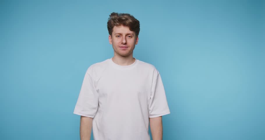A young adult stands confidently against a vibrant blue backdrop, flashing a thumbs-up and a bright smile. His casual white shirt complements his upbeat demeanor.