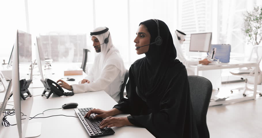 Young female Arabic call center employee in headset with mic talking to customer on audio call at computer monitor, typing on keyboard, giving technical support service, help