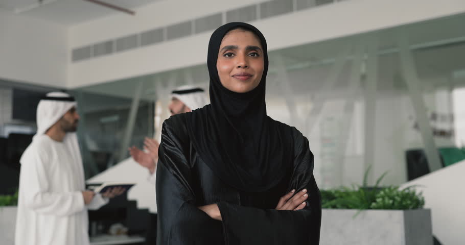 Happy confident young Muslim businesswoman standing in office with male colleagues in Arabic dresses working behind, keeping hands folded in confidence gesture, smiling for head shot portrait