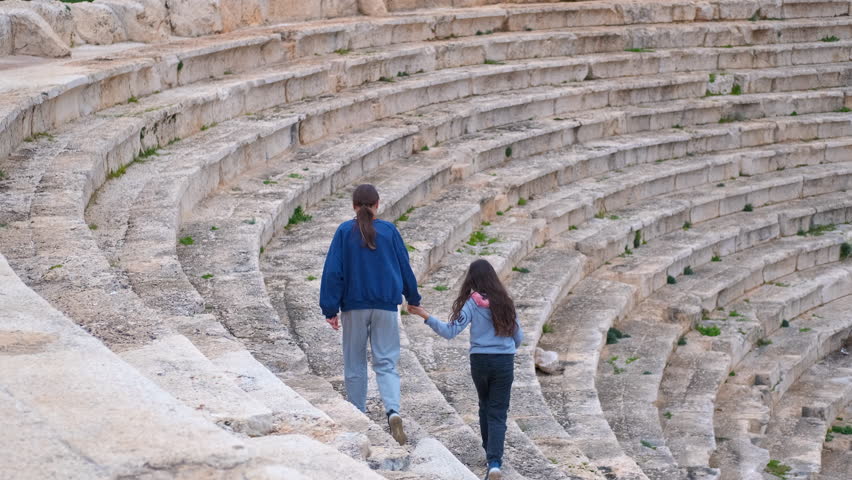 Tourists walking down ancient amphitheater stone steps. Young girls holding hands while descending weathered stone steps of ancient amphitheater, exploring historical ruins