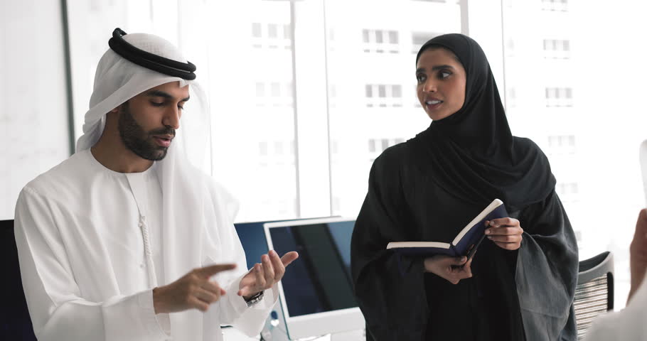 Business team of positive young Arabian professionals in traditional dresses talking in office, discussing teamwork, brainstorming on project together, sharing ideas, planning investment