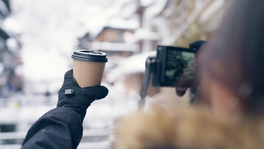 Happy young Asian woman drinking hot coffee and using mobile phone taking picture while travel Japan countryside Ginzan Onsen hot spring ryokan area in Yamagata prefecture on winter holiday vacation.