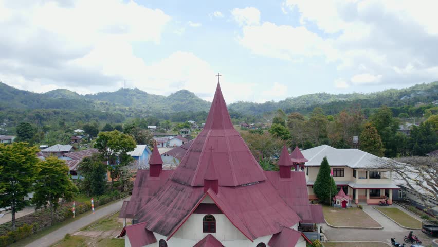 Aerial drone footage of a red roof church in Ruteng, Flores, Indonesia. The video captures the church architecture with a cross on top, set against a scenic mountain landscape and village houses.