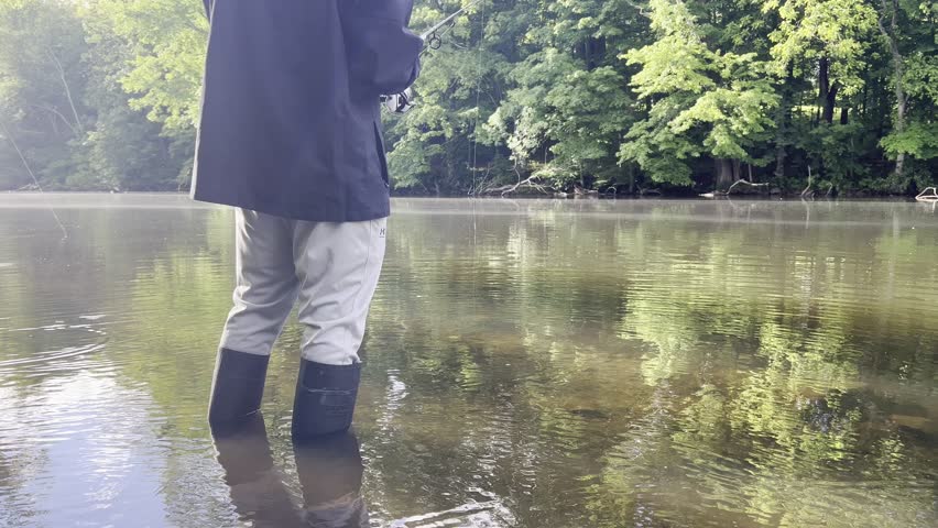 Man stands in shallow lake water wearing rubber boots, holding a fishing rod while casting. Peaceful outdoor fishing scene with calm water and natural surroundings.