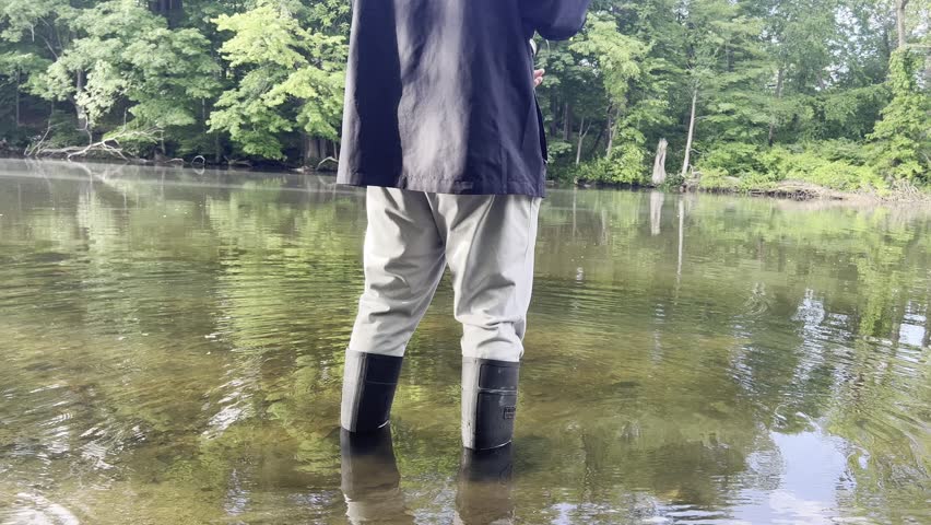 Man stands in shallow lake water wearing rubber boots, holding a fishing rod while casting. Peaceful outdoor fishing scene with calm water and natural surroundings.