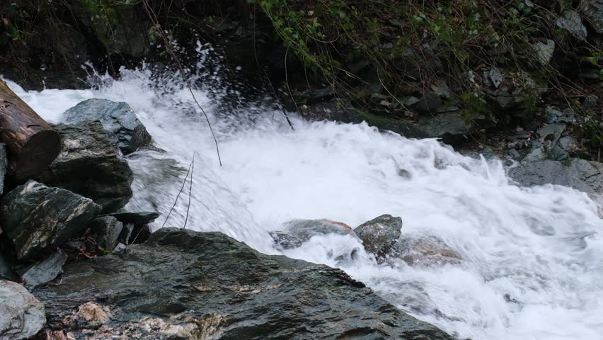 Water runs over rocks A log sits on the edge of the frame Green leaves are seen at the top The water is white as it flows quickly down hill