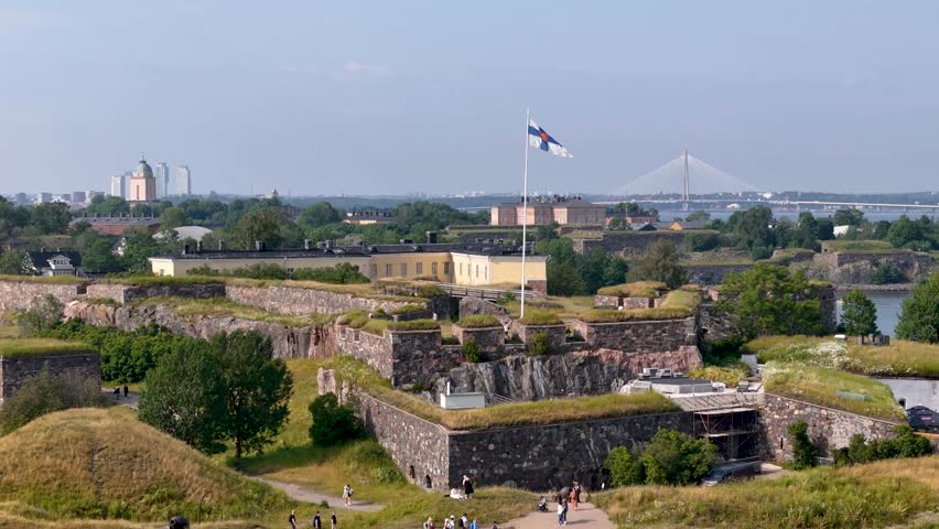 Slow, rising aerial view showing Finnish flag flying above the Suomenlinna sea fortress with city skyline behind it in Helsinki, Finland.