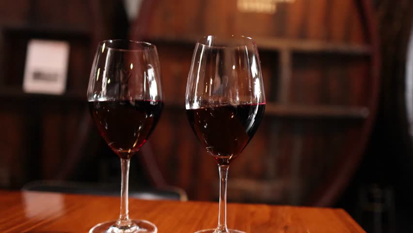 Two red wine glasses on wooden table, blurred person walking past, warm cellar lighting