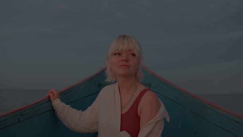 Young beautiful girl in red T-shirt and white blouse is sailing on boat along bay and enjoying local views smiling. Gray cloudy sky merges with surface of water on horizon