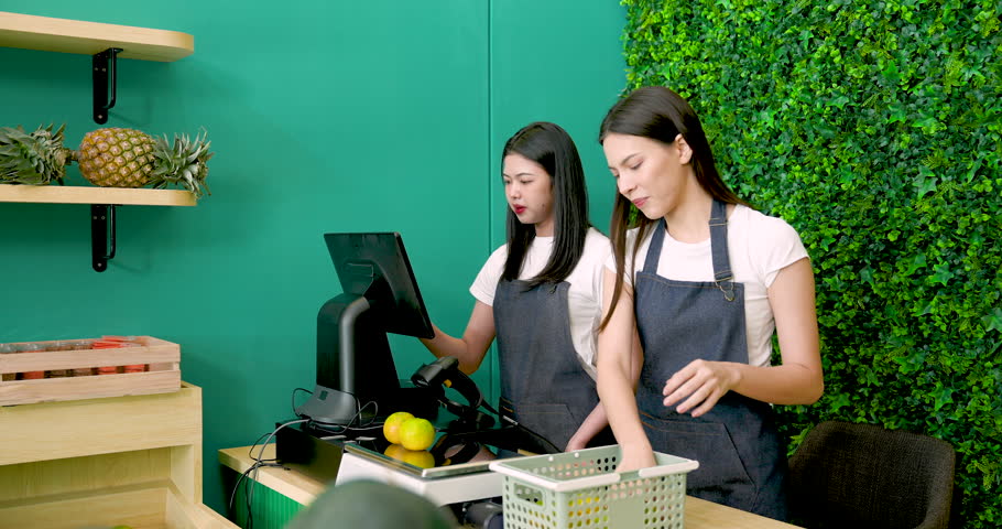 Asian female business owner using barcode scanner while Caucasian LGBTQ partner selects oranges from basket preparing fruit shop for opening with teamwork and care