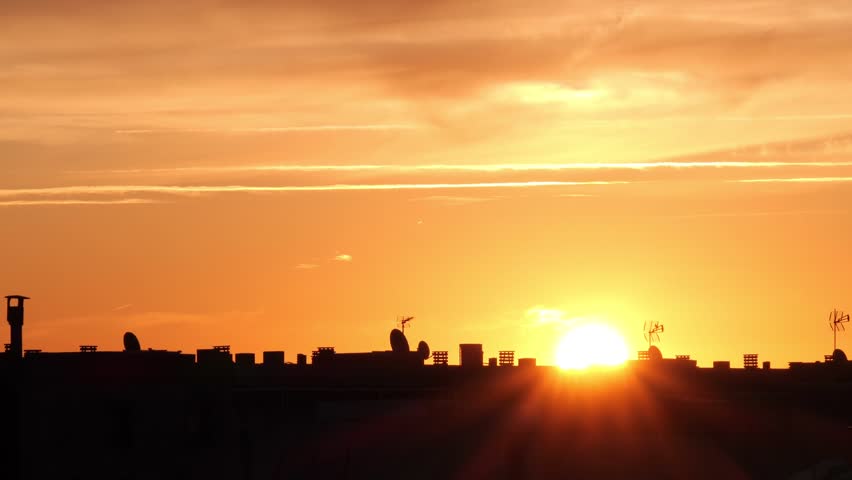 A vibrant sunset over a city skyline. The sun is low on the horizon, casting warm orange and yellow hues across the sky. Silhouettes of buildings are visible.