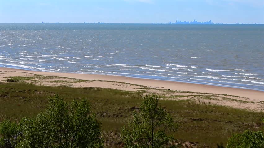 Gentle waves hit a sandy beach bordered by dune grasses with Chicago skyline on the horizon. Indiana Dunes National Park, USA
