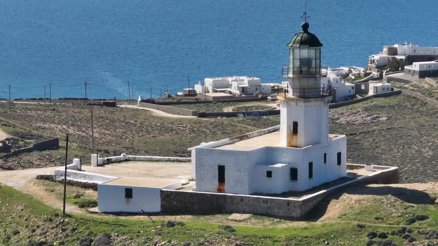 Drone pulls away, revealing a horizontal panoramic view of Armenistis Lighthouse in Mykonos, with the blue sea and sky creating a serene island atmosphere