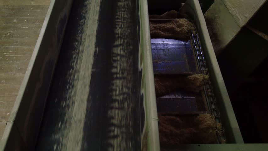 A close-up of industrial chain and belt conveyors transporting sawdust and wood fibers. This raw material, a byproduct of logging, is moved for recycling or further processing.