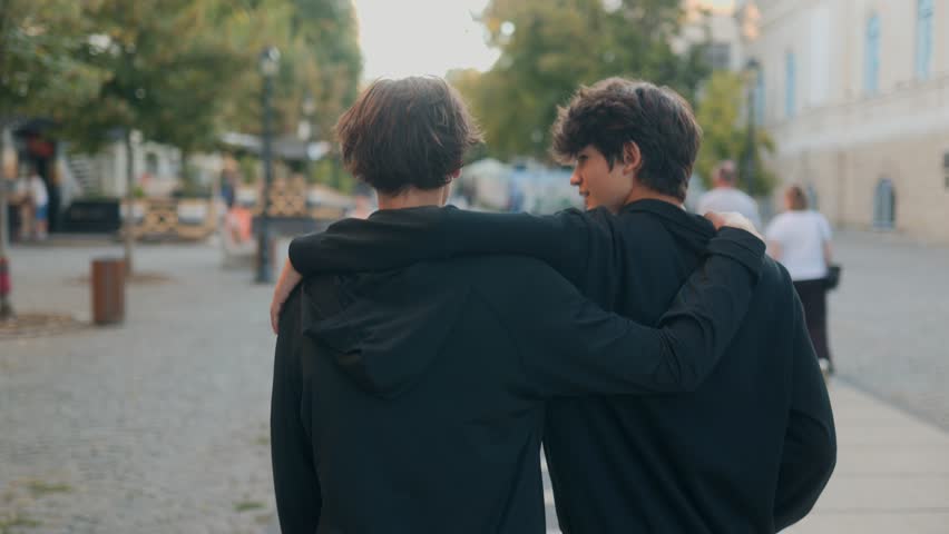 Smiling teen boys walk with arms on each other shoulders, showing closeness, trust, and carefree joy in an urban setting, highlighting their youthful lifestyle.
