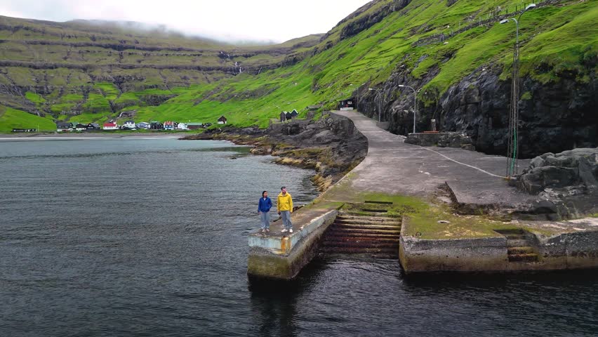 Two individuals stand on a stone pier by Tjornuviks calm waters, with lush hills and traditional Faroe Islands houses, embodying the beauty of this picturesque spot.