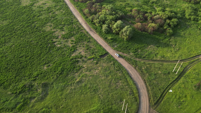 White car navigating dusty rural road, cutting through green fields and bordering forest with sweeping landscape view