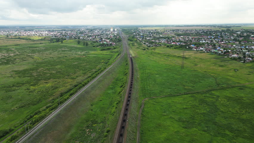 Freight train passing through countryside landscape, carrying goods across vast green fields towards a distant town under a cloudy sky