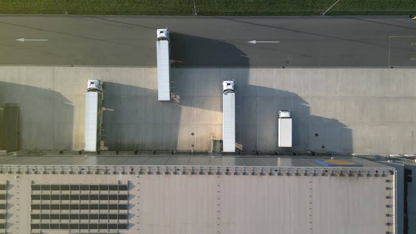 Aerial view of a modern distribution warehouse with loading docks. White cargo trucks are parked and maneuvering, showcasing efficient freight logistics and organized industrial operations.