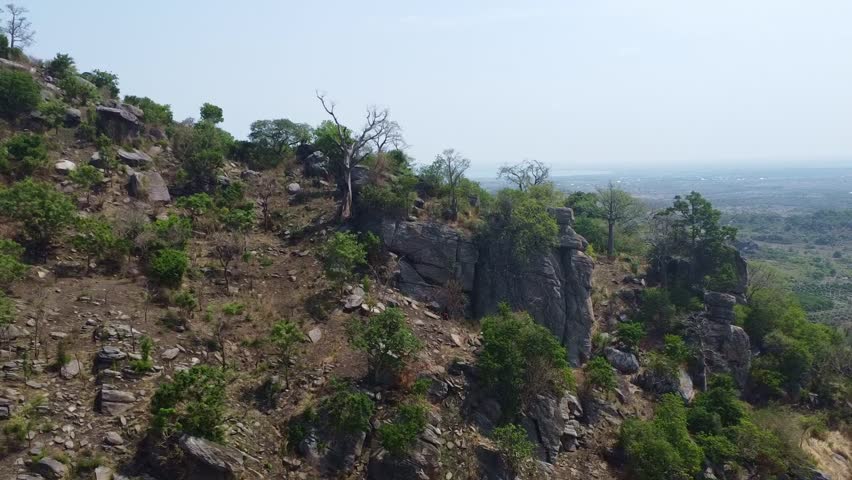 This wide-angle shot showcases a rocky hillside adorned with lush trees and vibrant shrubs. The landscape features distinct rock formations, with the arid plains stretching into the horizon under a cl