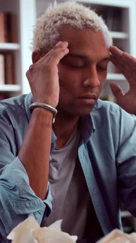 Stressed young African American creative professional sitting at cluttered desk holding his temples closing his eyes, overwhelmed by headache and paperwork, frustrated by writers block work pressure