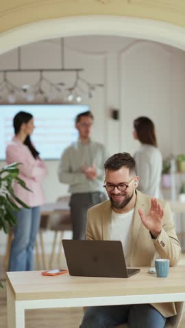 Focused businessman working on laptop, colleagues discussing in background, modern office environment