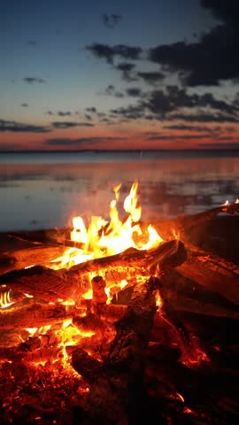 Warm flames dance on a bonfire built on a sandy beach, providing a mesmerizing spectacle against the backdrop of a tranquil sunset over a calm lake