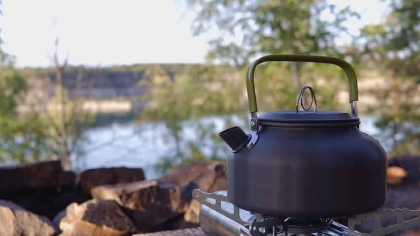 Kettle on a portable gas stove in a scenic forest landscape by the lake. Cozy camping atmosphere, outdoor cooking, and relaxation in nature.