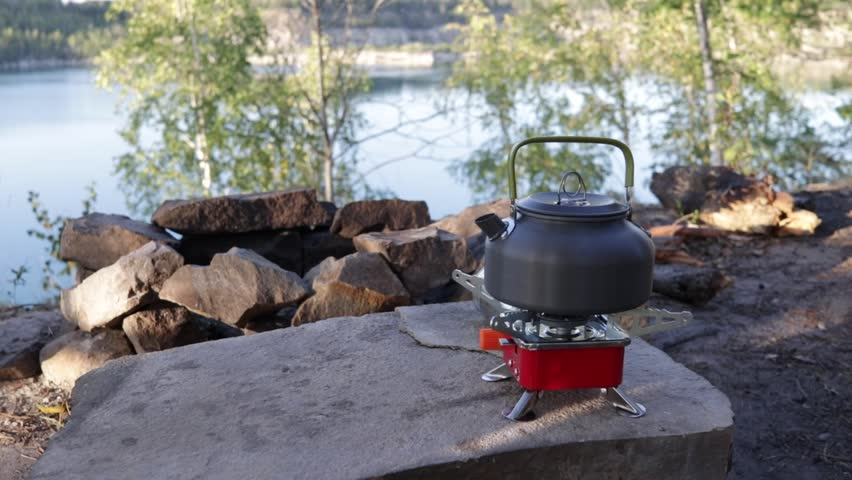 Kettle on a portable gas stove in a scenic forest landscape by the lake. Cozy camping atmosphere, outdoor cooking, and relaxation in nature.