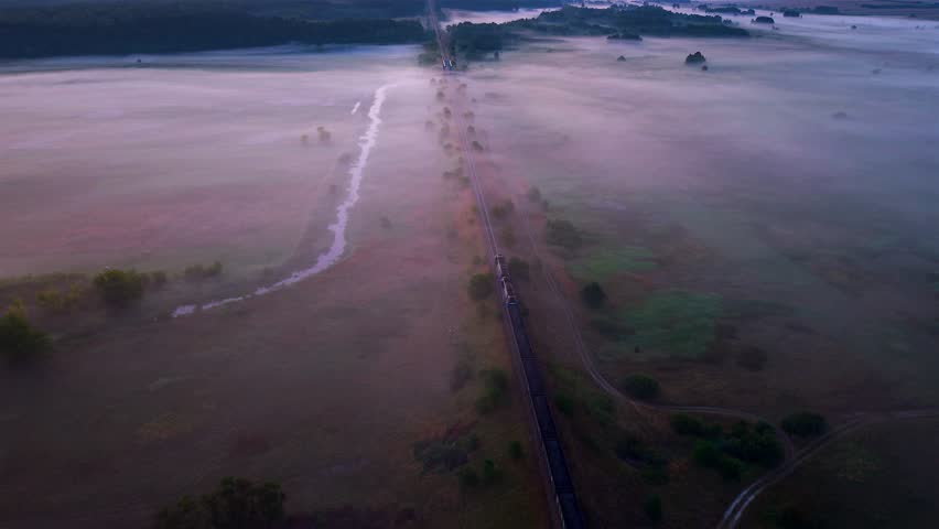 Aerial view, freight train moving along railway tracks covered in morning fog near the Nida River, Poland, with peaceful meadows and countryside landscape