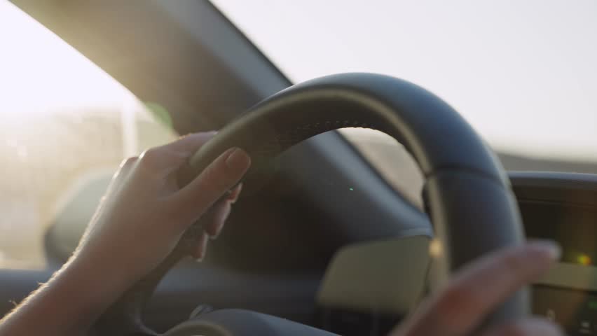 Female hands on car steering wheel driving on highway road with sun shining. Close up on wheel as woman holding it commuting to work or going on vacation or escaping. Sunset or sunrise in background