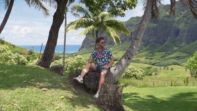 Tourist man in traditional Hawaiian shirt and flower lei relaxes under palm tree with scenic mountains and ocean view. Image represents vacation, aloha culture, USA tourism, famous Hawaiian islands - Powered by Shutterstock - Get 15% off with code: PIKWIZARD15