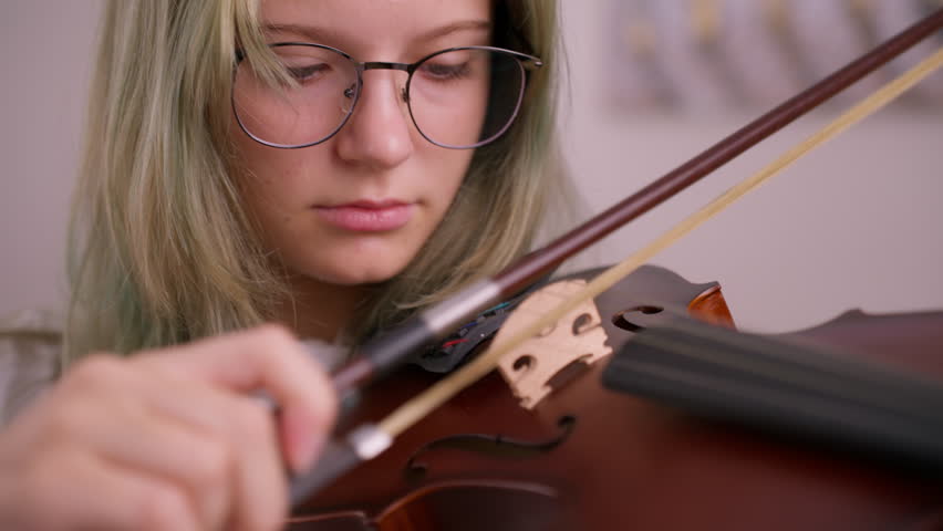 Teenager girl musician carefully adjusting violin bow, concentrating on instrument details before performance, displaying passion for classical music and artistic expression