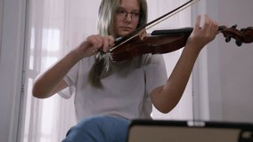 Talented young musician sits near a window, diligently practicing the violin with the help of an online lesson displayed on a tablet, showcasing dedication and passion for music education - Powered by Shutterstock - Get 15% off with code: PIKWIZARD15