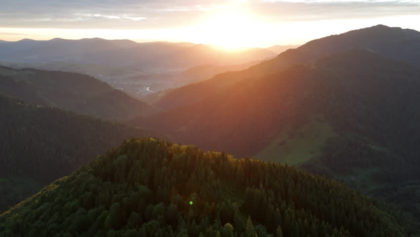 Aerial View of Sunrise Over Lush Green Mountain Landscape. Sun rises over misty forested mountains, casting warm light across lush green valleys and distant ridges.