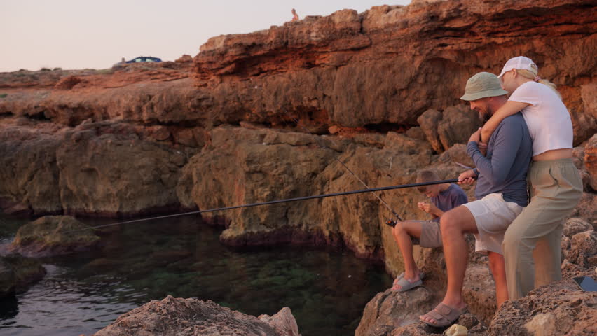 Family fishing together on rugged coastal terrain, parents guiding young son during warm sunset light, sharing outdoor bonding moment near rocky shoreline