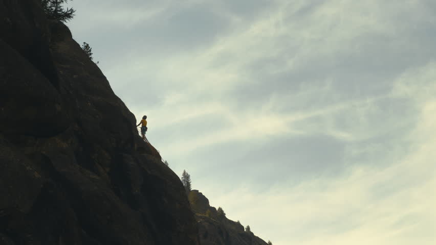 Silhouette of a climber scaling a rocky cliff with safety gear under cloudy sky, distant view