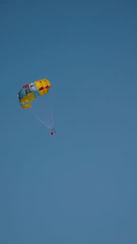 An unrecognizable person experiences the thrill of flying on a parachute with a colorful canopy, soaring against a clear sky, unrivaled adrenaline rush that will leave you in awe