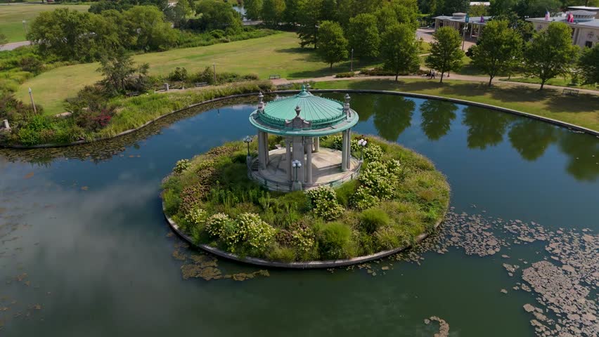 revolving aerial shot of a gazebo surrounded by a blue pond reflecting the sky and trees