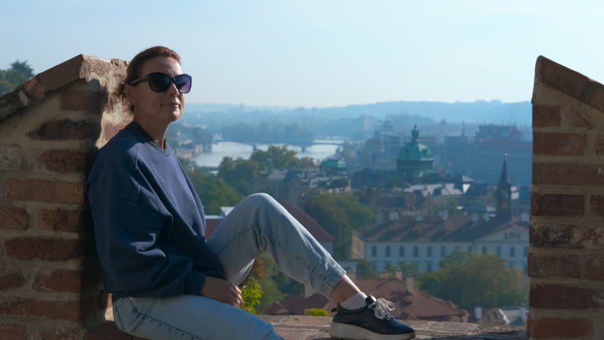Tourist enjoying panoramic view of prague from vysehrad fortress. Woman sitting on red brick wall, enjoying scenic prague cityscape, overlooking historical urban landscape with vltava river