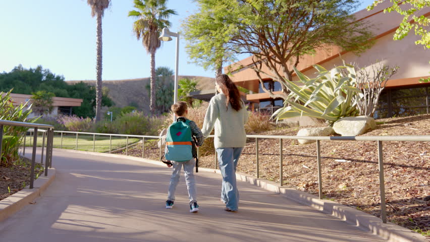 Loving mother and her cheerful child going to School holding each other hands along a sunlit pathway, surrounded by lush greenery and modern architecture that enhance their lovely environment