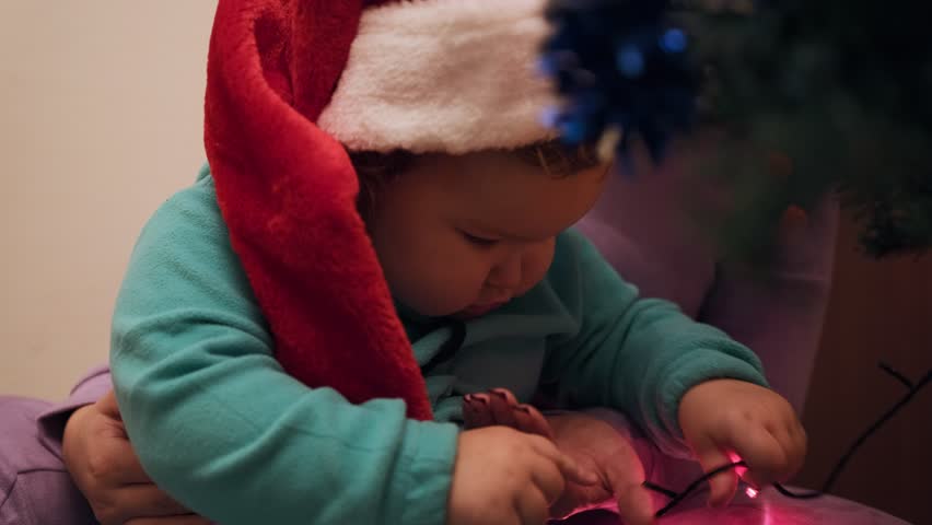 A young child wearing a Christmas hat reaches out to touch the colorful lights on the holiday tree, creating a joyful atmosphere at home. The spirit of the season is evident.