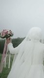 Back view of a bride wearing a white gown and veil, standing in a green field with a misty forest background, vertical video - Powered by Shutterstock - Get 15% off with code: PIKWIZARD15