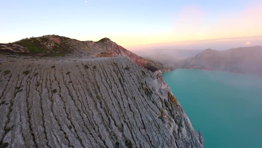 Cinematic FPV drone flight above rugged crater rim of Ijen Volcano at sunrise, overlooking turquoise sulfur lake in East Java, Indonesia. Stunning volcanic landscape and morning light.