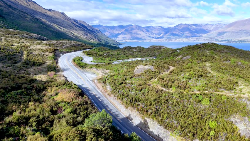 Drone glides above curving mountain road, lush greenery, and lake under bright daylight