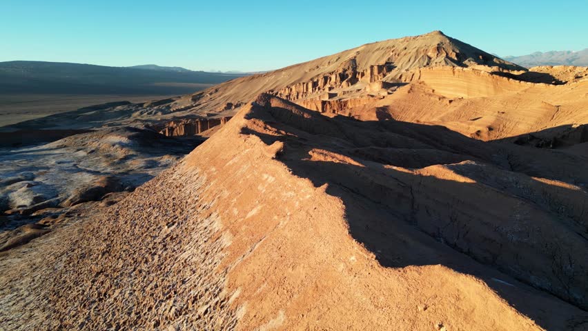 Drone aerial capturing the surreal desert of Valle de la Luna, with orange mountains glowing and salt flats shining at golden hour.