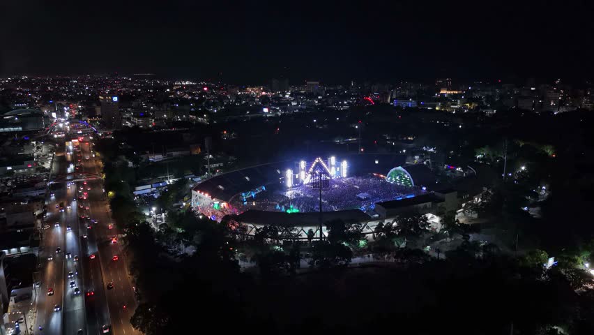 Wide establishing aerial drone shot of Barbarella music festival in a stadium at night, showing stage lights, large crowd, and city traffic, Dominican Republic