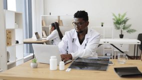 Male African healthcare professional analyzing X-ray results at desk in modern medical workspace. Scene conveys concept of medical analysis, professional healthcare duties, patient diagnostics - Powered by Shutterstock - Get 15% off with code: PIKWIZARD15