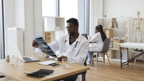 Male African healthcare professional analyzing X-ray results at desk in modern medical workspace. Scene conveys concept of medical analysis, professional healthcare duties, patient diagnostics - Powered by Shutterstock - Get 15% off with code: PIKWIZARD15