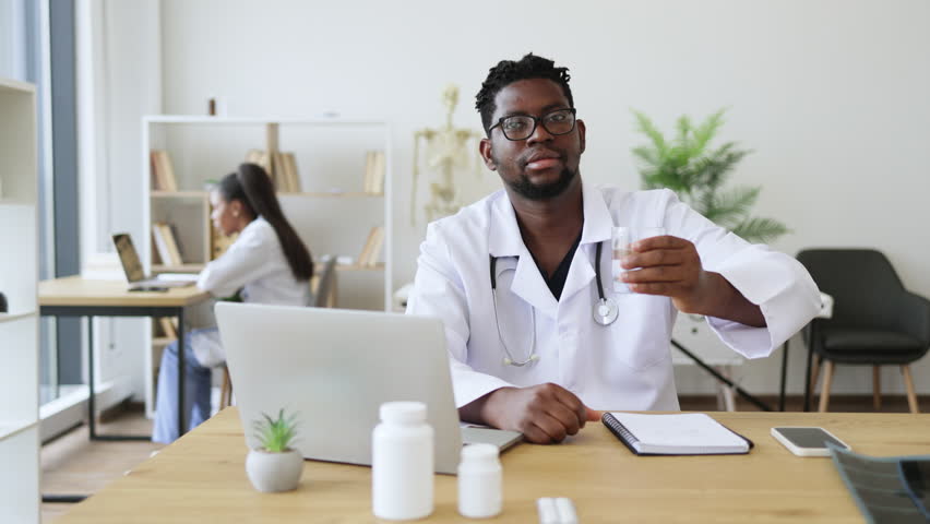 Male doctor in white coat holding glass of water, emphasizing drinking water benefits in health education within clinic environment. Demonstrates hydration importance, engages viewers on wellness